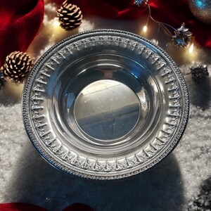 May include: A silver serving bowl with a decorative rim. The bowl is sitting on a white surface with red fabric and pine cones around it.