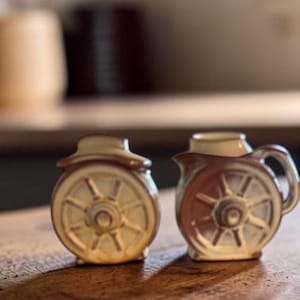 May include: A pair of ceramic salt and pepper shakers featuring a raised wagon wheel design on the front. The shakers are finished in a glossy glaze with cream and brown tones, and the right shaker includes a small handle and a pouring spout.