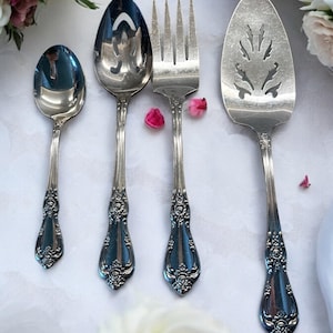 Puede incluir: Cuatro utensilios de servicio de plata con mangos adornados, que incluyen una cuchara ranurada, un tenedor de servicio, una cuchara y un cuchillo para pastel, sobre un mantel blanco.