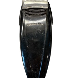 Puede incluir: Una afeitadora el&eacute;ctrica Remington negra con un protector de cuchilla de pl&aacute;stico transparente. La afeitadora tiene el nombre de la marca "Remington" impreso en la parte delantera. El cuerpo de la afeitadora es negro brillante.