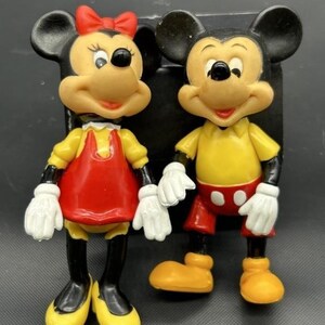 Pode incluir: Duas figuras vintage do Mickey Mouse e da Minnie Mouse. Mickey usa uma camisa amarela, cal&ccedil;&otilde;es vermelhos e sapatos amarelos. Minnie usa um vestido vermelho, sapatos amarelos e um la&ccedil;o vermelho. Ambos t&ecirc;m luvas brancas e orelhas pretas.