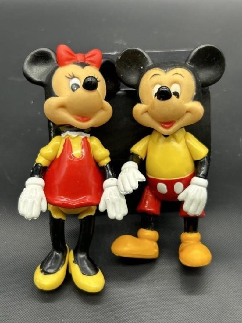 Pode incluir: Duas figuras vintage do Mickey Mouse e da Minnie Mouse. Mickey usa uma camisa amarela, cal&ccedil;&otilde;es vermelhos e sapatos amarelos. Minnie usa um vestido vermelho, sapatos amarelos e um la&ccedil;o vermelho. Ambos t&ecirc;m luvas brancas e orelhas pretas.