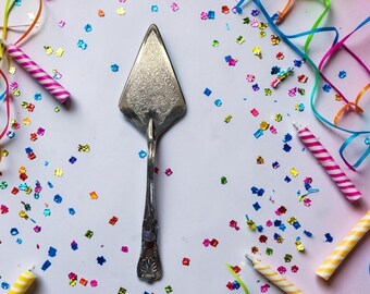 May include: A silver-toned metal cake server featuring an intricate, embossed floral pattern on the triangular blade and a decorative, shell-like motif at the base of the handle, positioned vertically against a white surface with confetti.