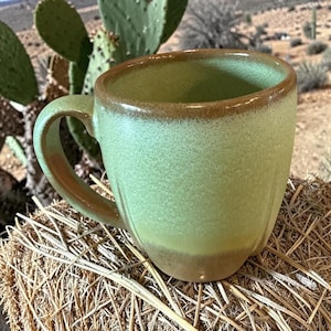 May include: A green ceramic mug with a brown rim and handle. The mug has a textured surface and is sitting on a bed of straw. The mug is a light green color with a darker green handle and a brown rim.