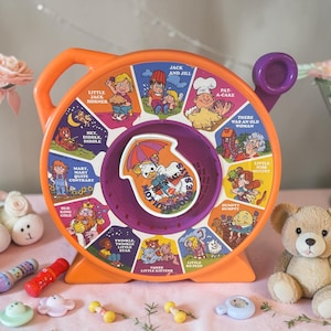 May include: An orange and purple vintage See 'N Say toy featuring illustrations of nursery rhymes. The toy has a handle and a dial with phrases like "Jack and Jill" and "Humpty Dumpty". A teddy bear and other toys are in the foreground.