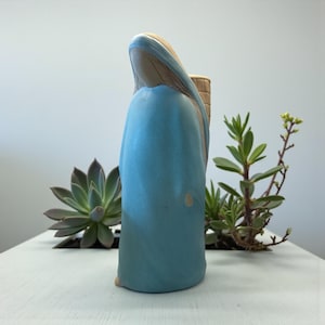 May include: A ceramic figurine of a woman in a blue robe with a tan head covering and a basket on her back. The figurine is on a white surface with green plants in the background.