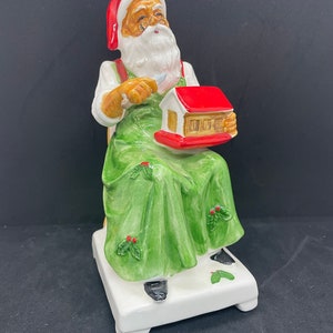 May include: A ceramic figurine of Santa Claus wearing a red hat and green overalls, sitting in a chair and holding a small red house. The figurine is decorated with holly leaves and berries.