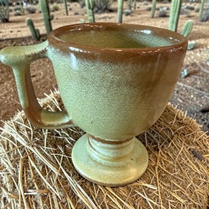 May include: A handmade ceramic mug with a pedestal base and a large handle. The mug has a green and brown glaze, with a textured surface. The mug is sitting on a bed of straw, with cacti in the background.