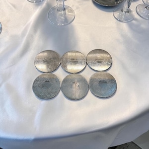 May include: A set of six circular silver-toned coasters arranged in two rows of three. Each coaster features a reflective metallic surface with a subtle, intricate etched pattern visible across the top, creating a uniform and polished appearance.