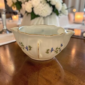 May include: A white porcelain gravy boat or sauce server with a scalloped rim and gold trim. The exterior features a delicate floral pattern of small blue flowers and green leaves. The vessel has a curved handle and a pouring spout on each side.