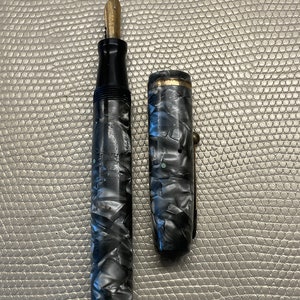 May include: A black and white marbled fountain pen with a gold-toned nib and cap. The pen is closed and the cap is off.