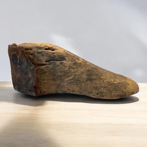 May include: A wooden shoe last, a tool used by shoemakers to shape shoes, with a worn, rustic appearance. The last is made of dark brown wood and has a metal piece on the heel.