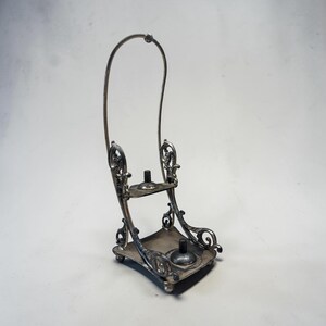 May include: An ornate silver-toned metal stand with a curved handle. The stand features two platforms and decorative scrollwork. The base is square with rounded corners and small feet. The stand is designed to hold two items.