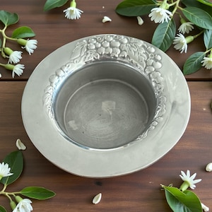 May include: A silver-colored, round dish with a decorative grape and leaf design around the rim. The dish has a shallow, concave center. The background is a dark wood surface with white flowers and green leaves.