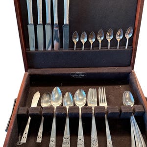 May include: A vintage silverware set displayed in a wooden case. The set includes knives, spoons, and forks with ornate handles. The case is lined with dark fabric, and the silverware is arranged neatly.