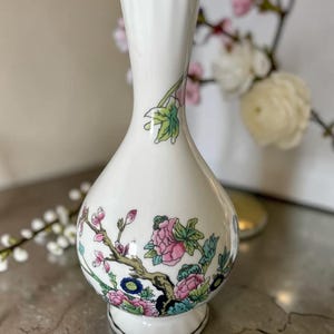 May include: A white porcelain vase with a narrow neck and a bulbous base, featuring a gold-trimmed rim and foot. The body is decorated with a colorful floral pattern of pink peonies, green leaves, and brown branches against a smooth white surface.