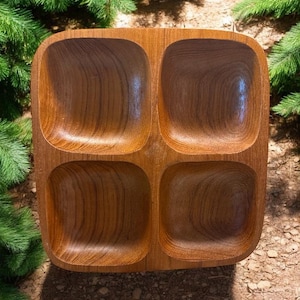 May include: A square, wooden serving tray with four compartments. The tray is made of light brown wood with visible grain patterns. The compartments are rounded squares, perfect for serving snacks or appetizers. The tray is set against a backdrop of greenery and earth.