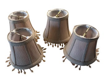 May include: Set of four beige lampshades with gold trim and fringe. The lampshades are pleated and have a delicate, feminine design.