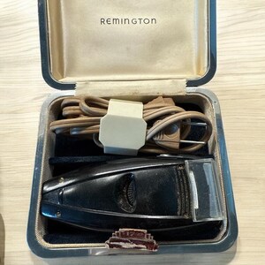 Puede incluir: Afeitadora el&eacute;ctrica Remington vintage en su estuche original. La afeitadora negra est&aacute; alojada en un estuche plateado con interior beige. El estuche contiene un cable de alimentaci&oacute;n y un peque&ntilde;o alfiler decorativo.