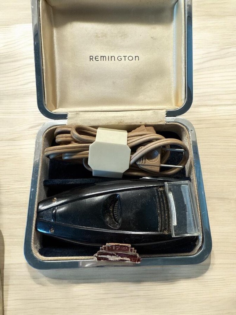 Puede incluir: Afeitadora el&eacute;ctrica Remington vintage en su estuche original. La afeitadora negra est&aacute; alojada en un estuche plateado con interior beige. El estuche contiene un cable de alimentaci&oacute;n y un peque&ntilde;o alfiler decorativo.
