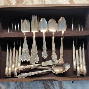 May include: A wooden box with a velvet lining holds a set of silverware. The set includes forks, knives, spoons, and a butter knife. The silverware is made of silver and has an ornate design.