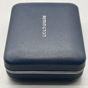 Puede incluir: Una caja rectangular azul marino con la palabra "Remington" impresa en blanco. La caja tiene una superficie texturizada y un borde plateado alrededor de los bordes. La caja est&aacute; cerrada y parece estar hecha de un material duradero.