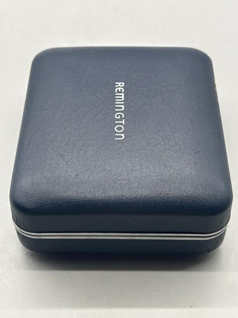Puede incluir: Una caja rectangular azul marino con la palabra "Remington" impresa en blanco. La caja tiene una superficie texturizada y un borde plateado alrededor de los bordes. La caja est&aacute; cerrada y parece estar hecha de un material duradero.
