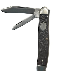 May include: A folding pocket knife with two open steel blades and a dark, textured handle. The handle features a small, silver-colored shield-shaped inlay and three metal pins. The metal bolster is smooth and rounded at the top and bottom ends.