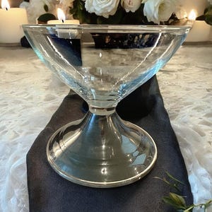 May include: A clear glass bowl with a wide, triangular top and a silver-colored base. The bowl sits on a dark blue cloth, with white flowers and lit candles in the background. The bowl is designed for serving.