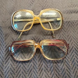 May include: Two pairs of eyeglasses with clear lenses. The top pair has a light brown frame with a rectangular shape. The bottom pair has a brown and amber frame with a rectangular shape.