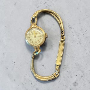 May include: Vintage gold-toned Bulova wristwatch with a round face and a textured band. The watch face features gold numerals and hands. The band is a woven style with a rectangular clasp.