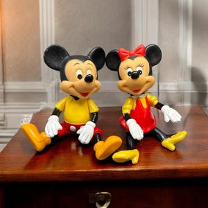Pode incluir: Duas estatuetas de Mickey e Minnie Mouse. Mickey usa uma camisa amarela, cal&ccedil;&otilde;es vermelhos e sapatos amarelos. Minnie usa um vestido vermelho, sapatos amarelos e um la&ccedil;o vermelho. Ambos t&ecirc;m orelhas pretas e luvas brancas. Est&atilde;o sentados numa superf&iacute;cie de madeira.