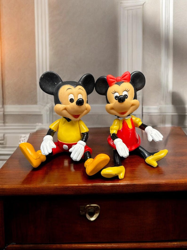 Pode incluir: Duas estatuetas de Mickey e Minnie Mouse. Mickey usa uma camisa amarela, cal&ccedil;&otilde;es vermelhos e sapatos amarelos. Minnie usa um vestido vermelho, sapatos amarelos e um la&ccedil;o vermelho. Ambos t&ecirc;m orelhas pretas e luvas brancas. Est&atilde;o sentados numa superf&iacute;cie de madeira.