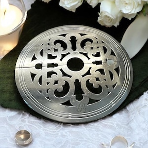May include: A silver metal floral design trivet with a central hole and decorative cutouts. The trivet is round and sits on a white lace surface. A lit candle and white roses are in the background.