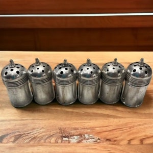 May include: Six silver metal salt and pepper shakers with a hole-punched top and a rounded bottom. The shakers are lined up in a row on a wooden surface.