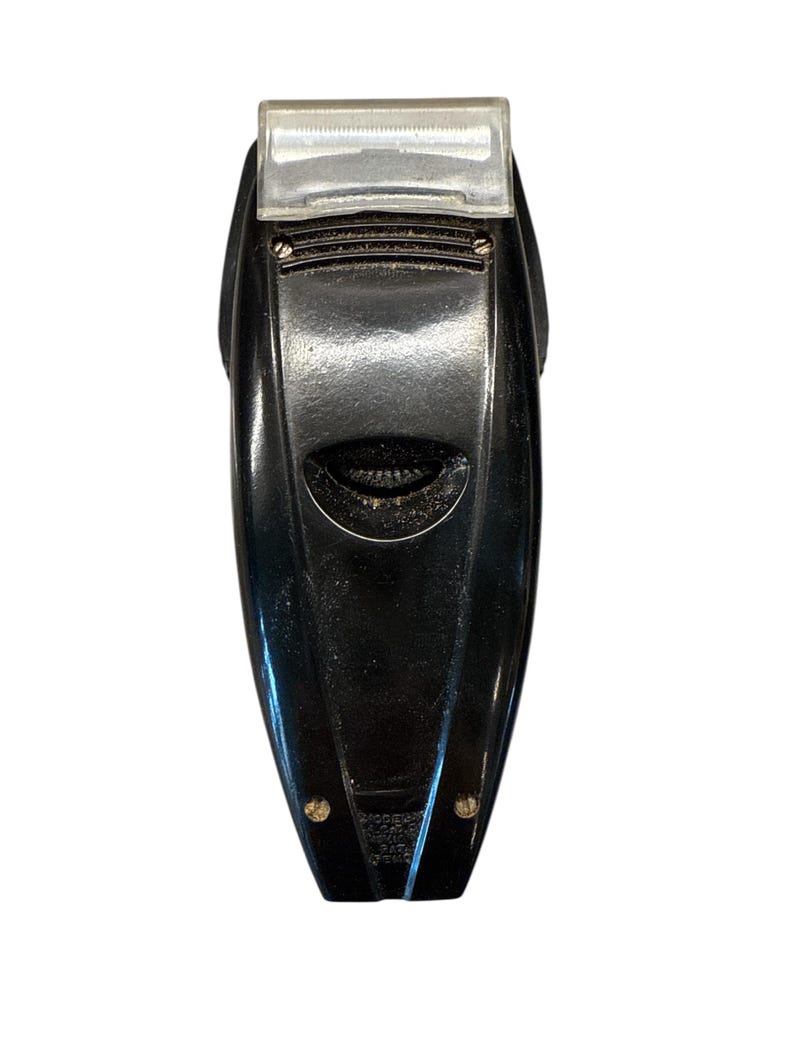 Puede incluir: Una afeitadora el&eacute;ctrica negra vintage con una cubierta protectora de pl&aacute;stico transparente. La afeitadora tiene un dise&ntilde;o aerodin&aacute;mico con un agarre texturizado y dos peque&ntilde;os detalles dorados en la parte inferior.