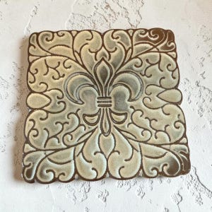May include: Square ceramic coaster with a fleur-de-lis design. The coaster features a light green and brown color scheme with a detailed, ornate pattern. The edges are a darker brown, creating a framed effect.