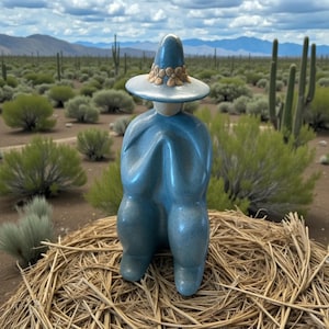 May include: A blue ceramic sculpture of a seated figure wearing a hat with a decorative band. The figure is positioned on a bed of straw, with a desert landscape in the background featuring cacti and mountains.