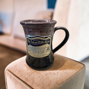 May include: A dark ceramic mug with a flared rim and a speckled purple and black glaze. The front features a cream-colored label with an illustration of a building and the text: The Inn at Ellis River, Jackson, New Hampshire, innatellisriver.com.