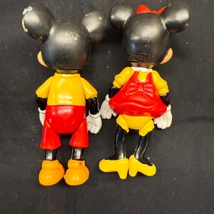 Pode incluir: Duas figuras vintage do Mickey e da Minnie Mouse. Mickey usa uma camisa amarela, cal&ccedil;&otilde;es vermelhos e sapatos laranja. Minnie usa um vestido amarelo com la&ccedil;o e saia vermelhos, e um la&ccedil;o vermelho na cabe&ccedil;a. Ambos t&ecirc;m orelhas pretas e luvas brancas.