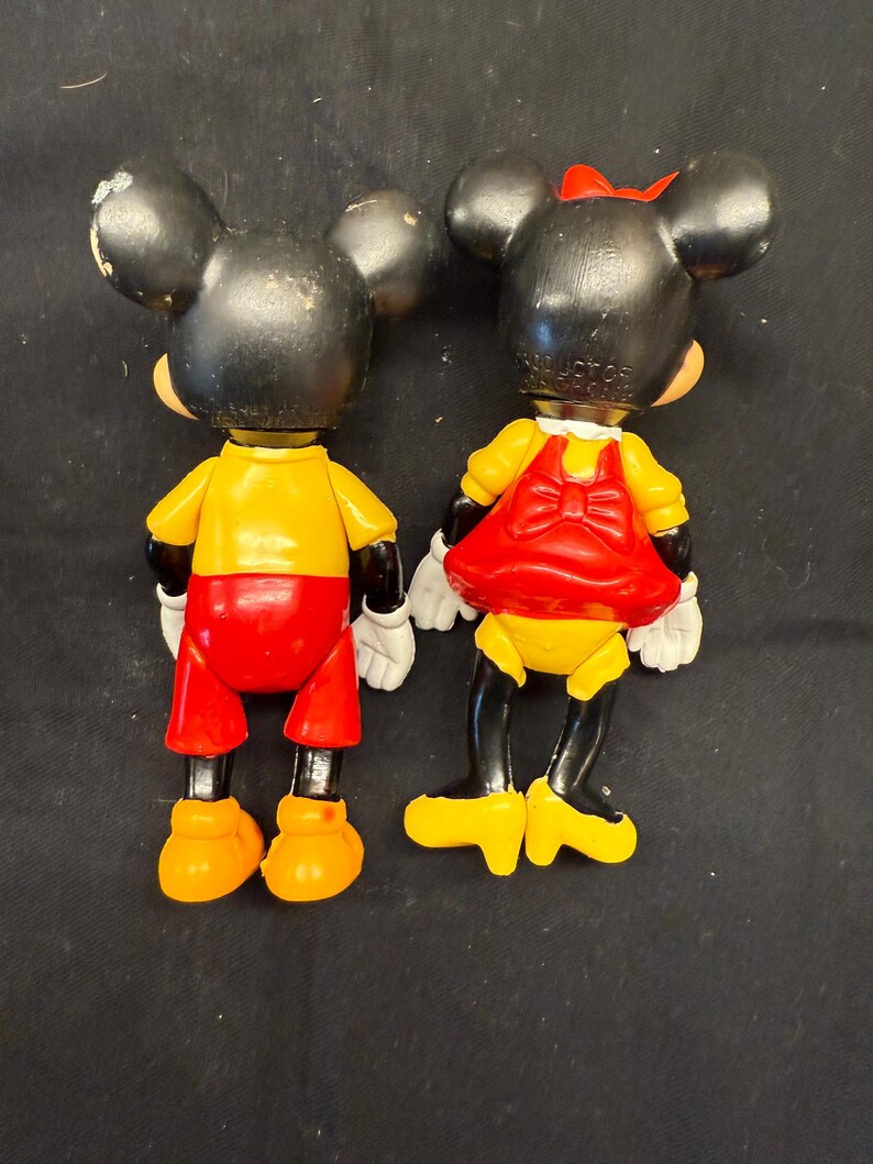 Pode incluir: Duas figuras vintage do Mickey e da Minnie Mouse. Mickey usa uma camisa amarela, cal&ccedil;&otilde;es vermelhos e sapatos laranja. Minnie usa um vestido amarelo com la&ccedil;o e saia vermelhos, e um la&ccedil;o vermelho na cabe&ccedil;a. Ambos t&ecirc;m orelhas pretas e luvas brancas.