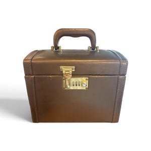 May include: A metallic bronze vanity case with a structured rectangular shape, a top handle, and a gold-toned combination lock mechanism on the front. The exterior features smooth surfaces with stitched detailing along the edges and corners.