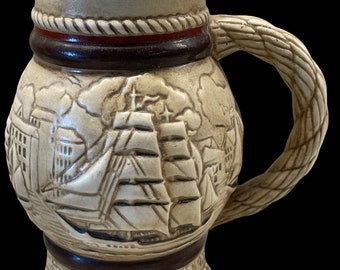 May include: A ceramic beer stein with a rope-like handle and a detailed image of a sailing ship. The stein has a brown band with a red rope design around the top and bottom. The text "Eine Bark St. Stausoi" is printed on the bottom of the stein.