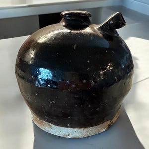 May include: A dark brown, glazed ceramic jug with a rounded body, a spout, and a small opening at the top. The jug has a glossy finish and a light-colored base. The design is simple and functional, suitable for holding liquids.