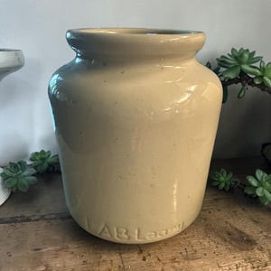 May include: A beige ceramic jar with a rounded body and a slightly flared rim. The jar has a smooth, glossy finish and the text "LAB Lagell" is visible near the base. The jar is likely vintage or antique.