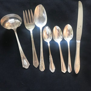May include: An assortment of vintage silverware, including a ladle, fork, spoons of various sizes, and a knife. The silverware is silver-toned and arranged on a dark background. The utensils have decorative handle designs.