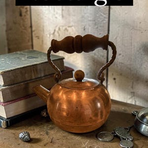 May include: A copper tea kettle with a rounded body, a short spout, and a lid featuring a small knob. The kettle has a twisted metal handle topped with a dark brown, turned wooden grip. The word Nostalgia is printed in white on a black bar.