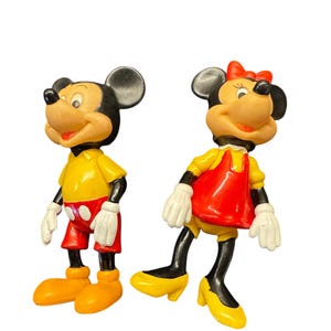 Pode incluir: Duas figuras vintage do Mickey Mouse e da Minnie Mouse. Mickey usa uma camisa amarela, cal&ccedil;&otilde;es vermelhos e sapatos laranja. Minnie usa um vestido vermelho, sapatos amarelos e um la&ccedil;o vermelho. Ambos t&ecirc;m luvas brancas e orelhas pretas.