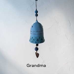 May include: A light blue ceramic bell with the inscription "I hope you know how real love means it." The bell has a decorative pattern at the bottom and is suspended by a string with blue beads. A small metal heart hangs below. The word "Grandma" is at the bottom.