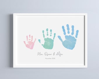 Könnte beinhalten: Ein weißer gerahmter Druck mit drei Handabdrücken in Aquarell. Die Handabdrücke sind in Rosa-, Grün- und Blautönen gehalten. Der Text unter den Handabdrücken lautet "Mia, Oscar & Alfie, November 2022".